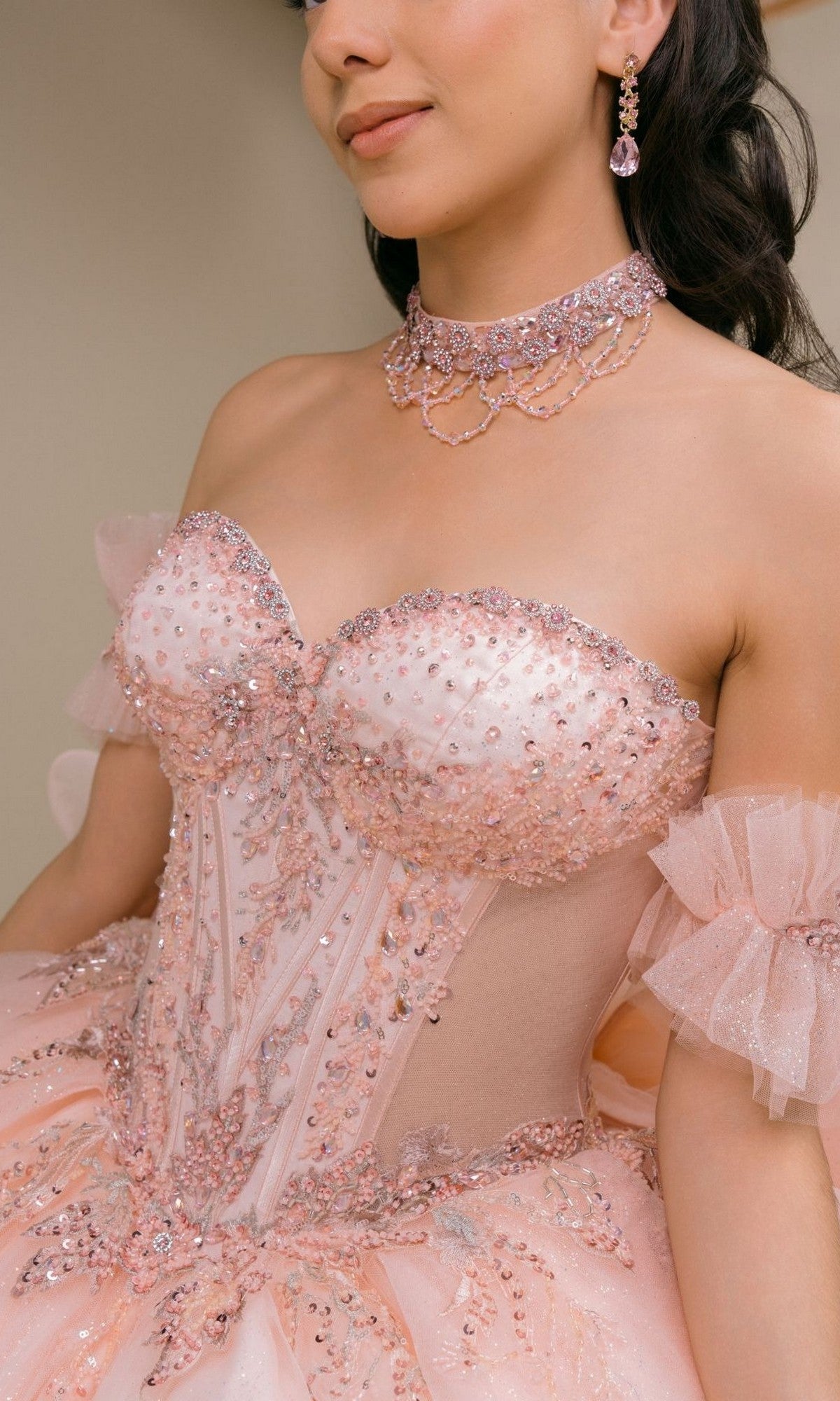 A woman in the Quinceanera Dress 8540JQ by Cinderella Coutureโa blush pink beaded gown with sheer panelsโstands against a neutral background, accessorized with a matching choker and earrings.